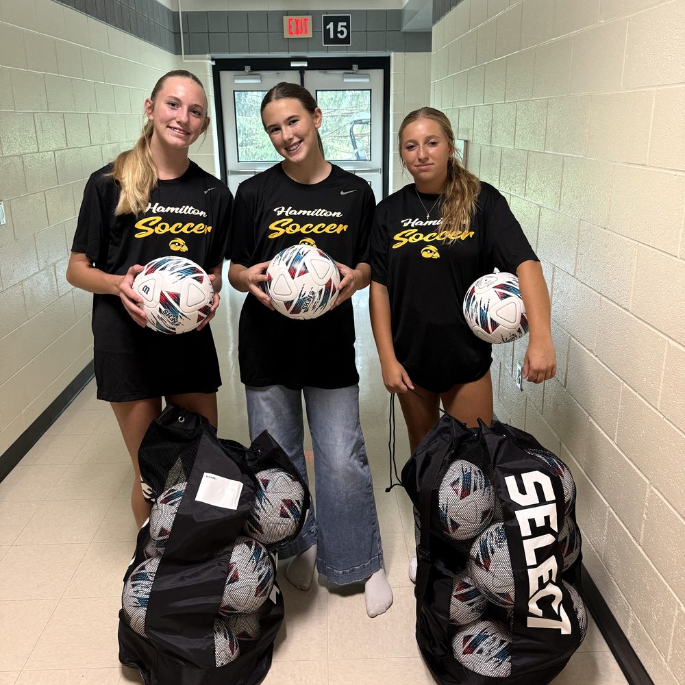 Three Hamilton girls soccer players holding new match balls, with two full ball bags at their feet